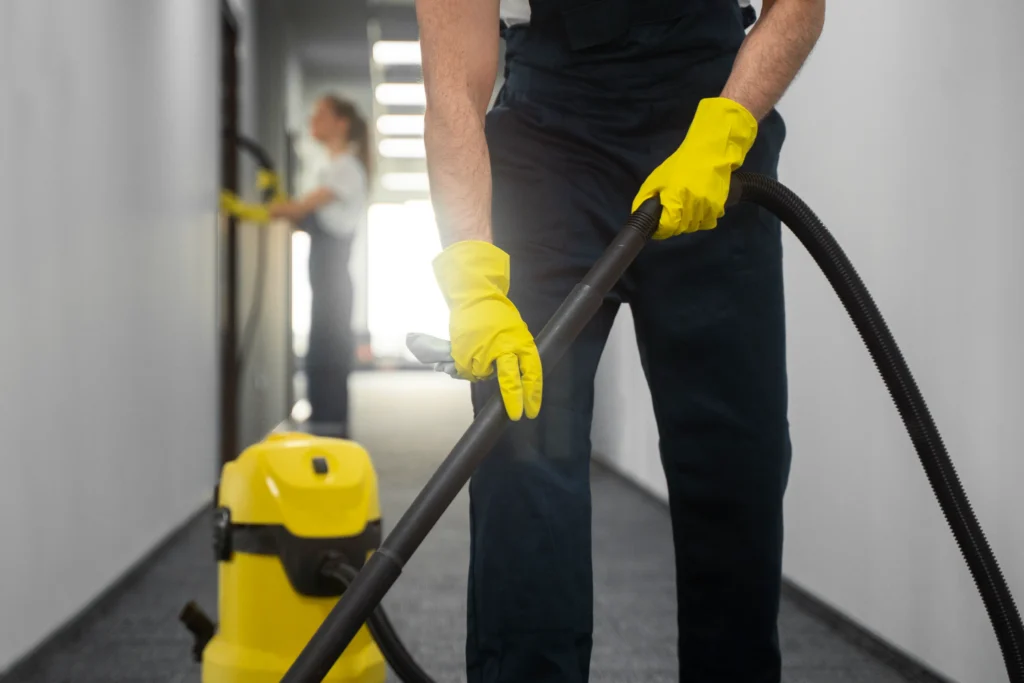 Commercial cleaner in yellow gloves using vacuum hose in modern white hallway - professional cleaning services